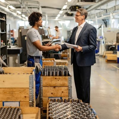 Happy quality control inspector shaking hands with African American steel worker while visiting industrial facility.