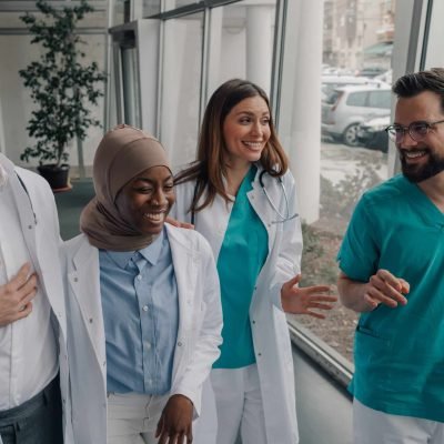 Four cheerful medical professionals, including a muslim woman wearing hijab, are walking and laughing together in a modern hospital corridor, enjoying a moment of camaraderie and positive energy