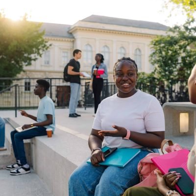 Portrait of intelligent multicultural university student girl sitting on staircase at college campus with her classmate and explaining lectures during coffee break. In background are students talking.